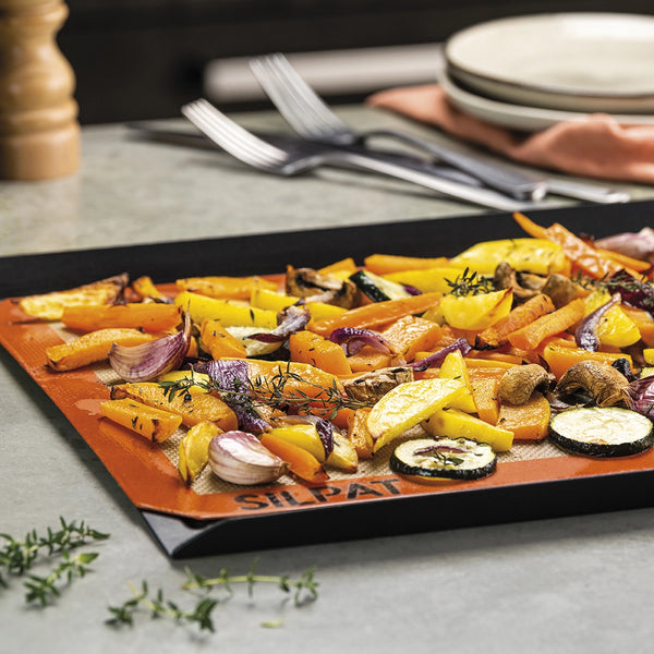 Roasted vegetables on a Silpat baking mat with a blurred background of plates and cutlery.